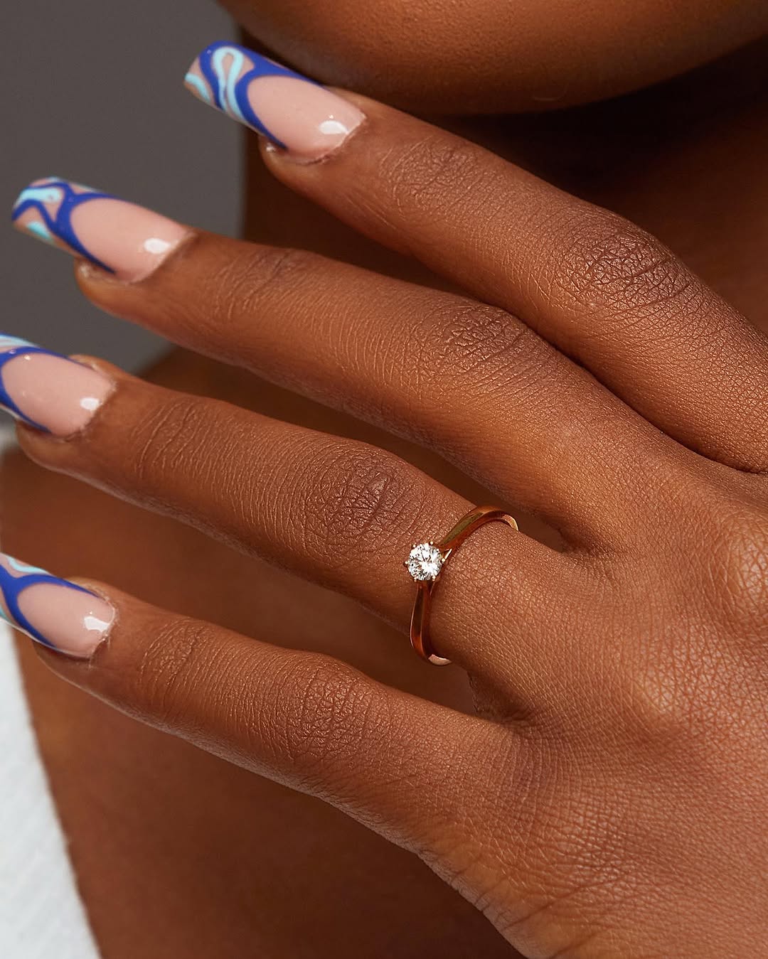 A person's hand with a natural diamond ring on their finger, alongside a manicured nail with blue and white stripes.