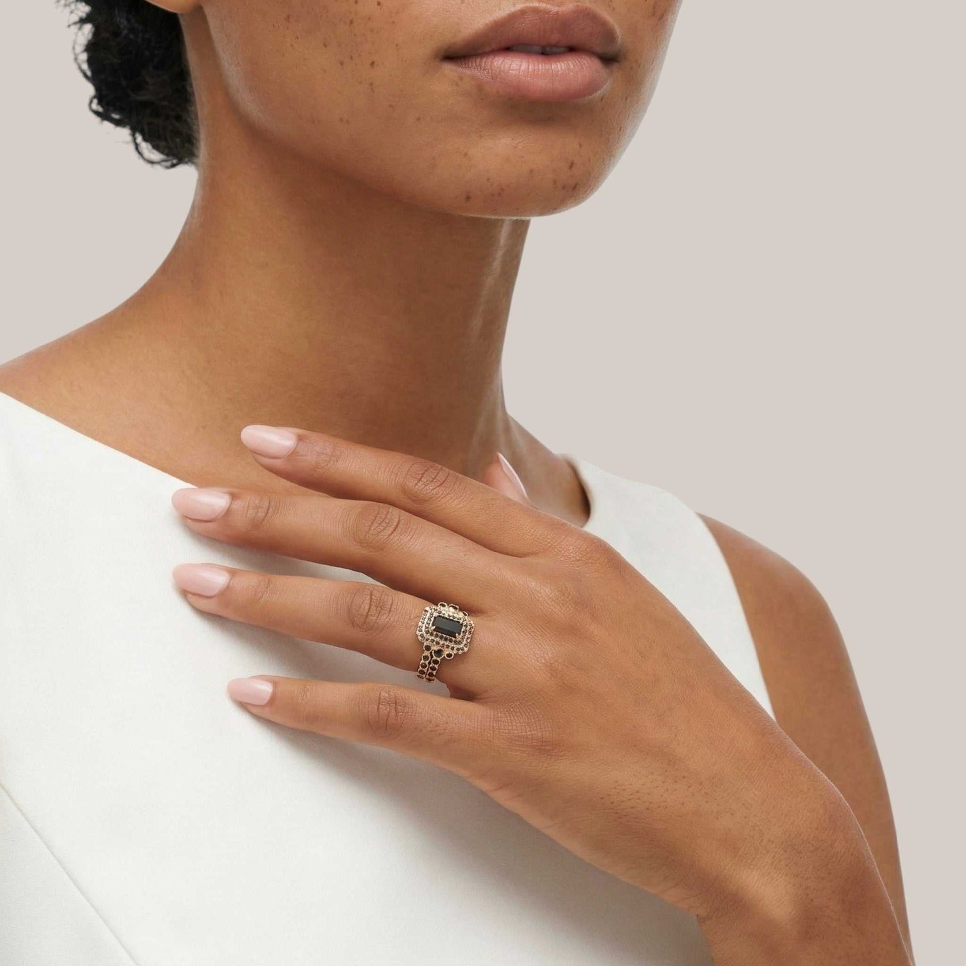 Close-up of a hand wearing a ring with a green gemstone, touching the shoulder against a neutral background.