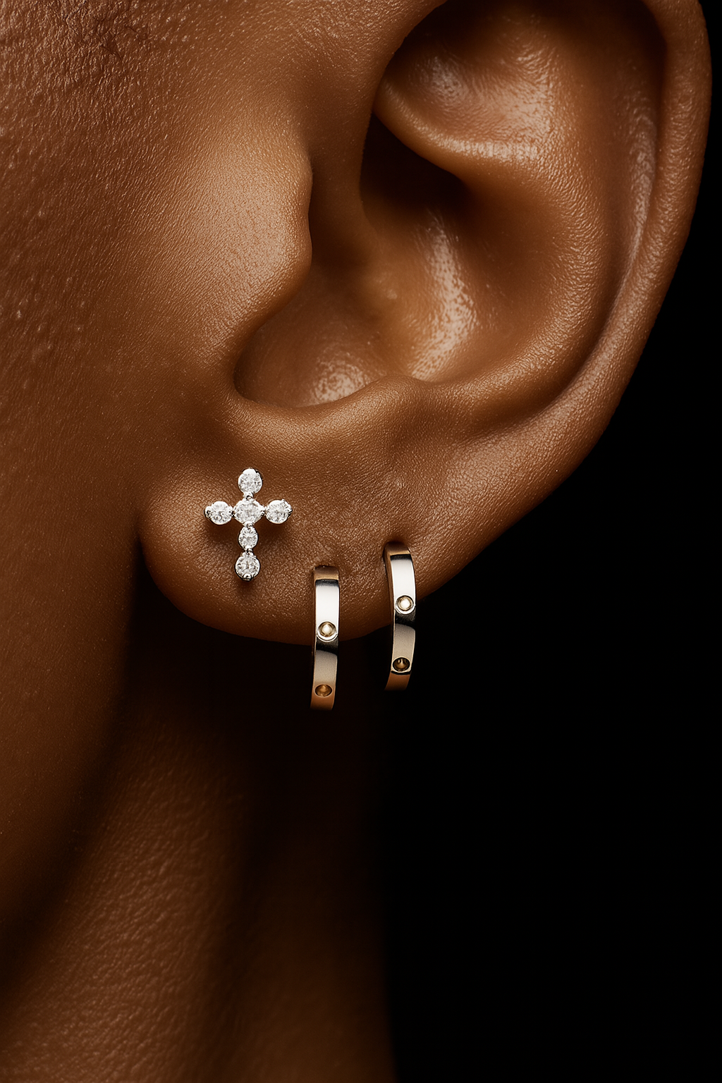Close-up of an ear wearing a diamond cross earring with two small hoop earrings.